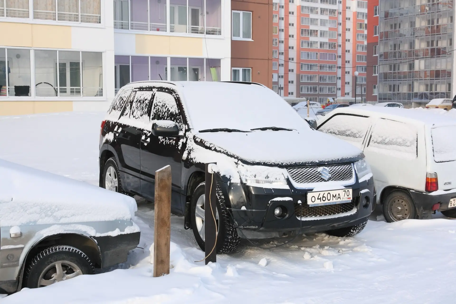 A snow-covered SUV parked outside an apartment block with a power cord running from a wooden post to its front end